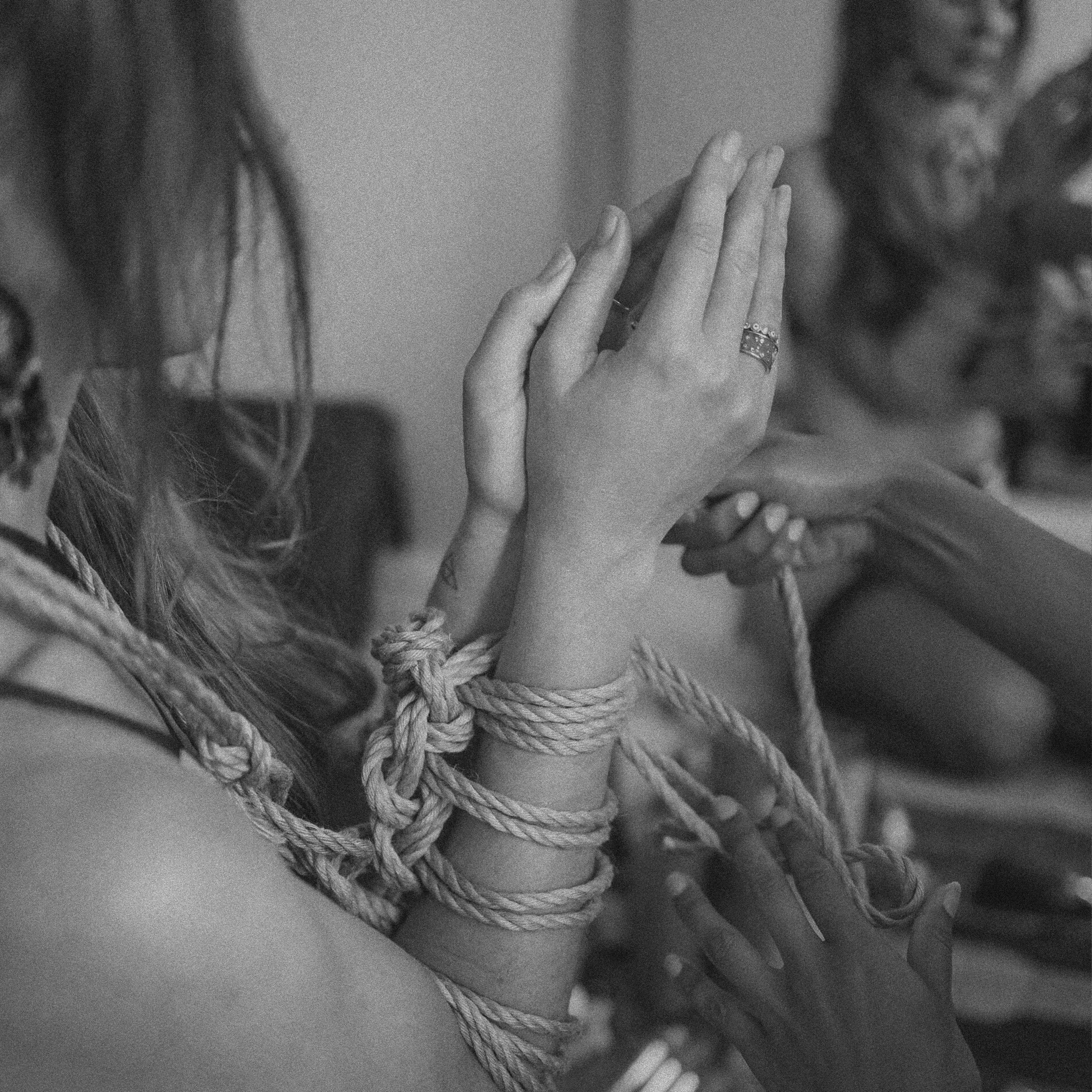 Close-up of a person’s hands wrapped in a rope, with a ring on one finger, and blurred people in the background.