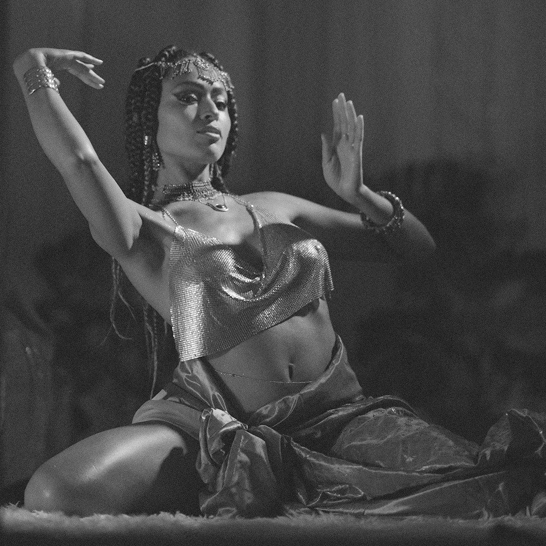A woman in a metallic top and loose pants poses with one arm raised and the other bent, wearing jewelry and an elaborate headpiece, in a black and white photo.