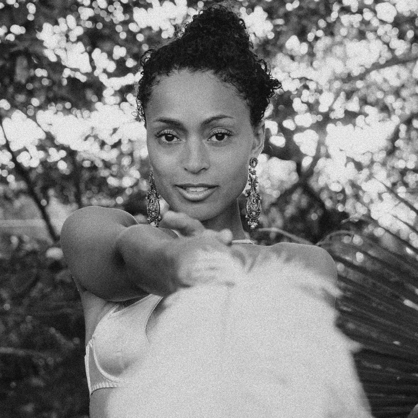 A black and white photograph of a woman with curly hair styled in an updo, wearing large, ornate earrings, and looking directly at the camera with a gentle expression. She is surrounded by leafy plants, and her arm is extended towards the camera, partially blocking her face.